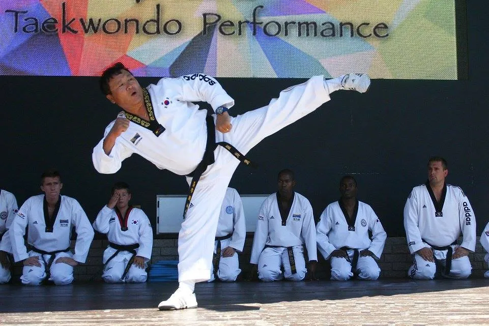 Grandmaster Eun Woo Lee performing a side kick at a Taekwondo performance, students seated behind him.